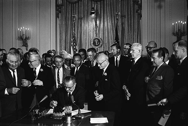 A black and white caption of President Lyndon B. Johnson signing the Civil Rights Act of 1964. Johnson is seated at a large desk, with numerous Congressmen and other men surrounding him. Dr. Martin Luther King stands directly behind him.