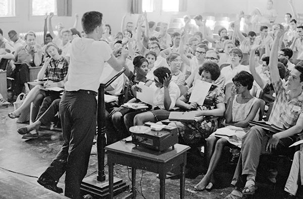 A black and white photograph of Dr. Staughton Lynd lecturing to a group of Freedom School teachers. Lynd stands at a podium in front of the group in a white t-shirt and black pants. Some members of the group have their hands raised. 