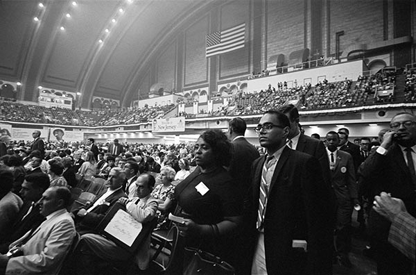 A black and white photograph of the Democratic National Convention in Atlantic City, New Jersey. Fannie Lou Hamer and Robert (Bob) Moses lead a group of Freedom Democratic Party members down the center aisle. 