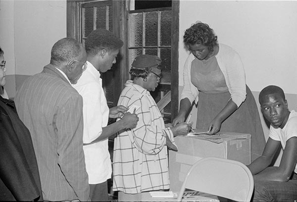 A black and white photograph of people standing in line to cast their Freedom Vote at a masonic temple. There are four people in line. A woman stands behind the ballot box, and a man is seated next to her.