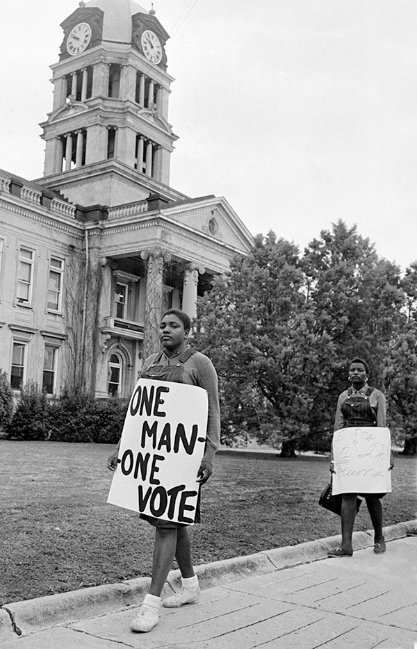 A black and white photograph of two Black protestors outside of the Leflore County courthouse. Both women are dressed in overall dresses with long sleeve shirts underneath. The woman in front holds a sign that reads: “One Man – One Vote.”