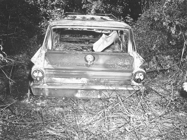 A black and white photograph of the burned car that James Chaney, Andrew Goodman, and Michael Schwerner drove to investigate the church burning. The car is surrounded by trees and debris. 