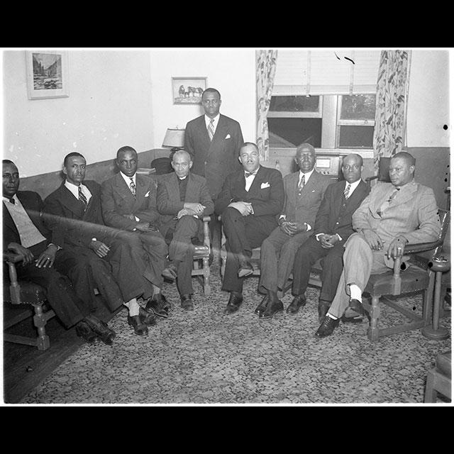A group of nine Black men sit together and look directly at the camera