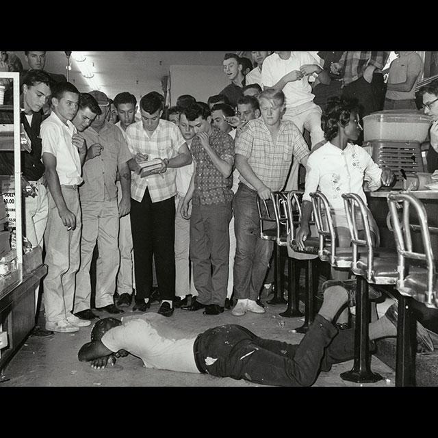 A Black protestor lies on the floor, covered in debris at the 1963 Woolworth's sit-in.